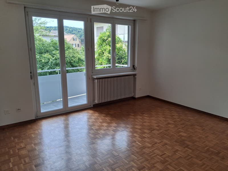 Empty room with wooden parquet flooring, large windows, radiator, and sliding glass door leading to a balcony.