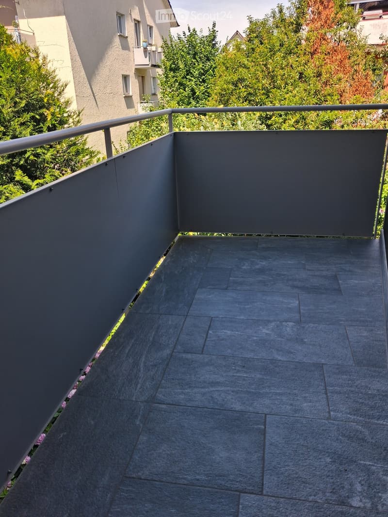 Modern balcony with grey tiled flooring, metal railing, and lush greenery in the background.
