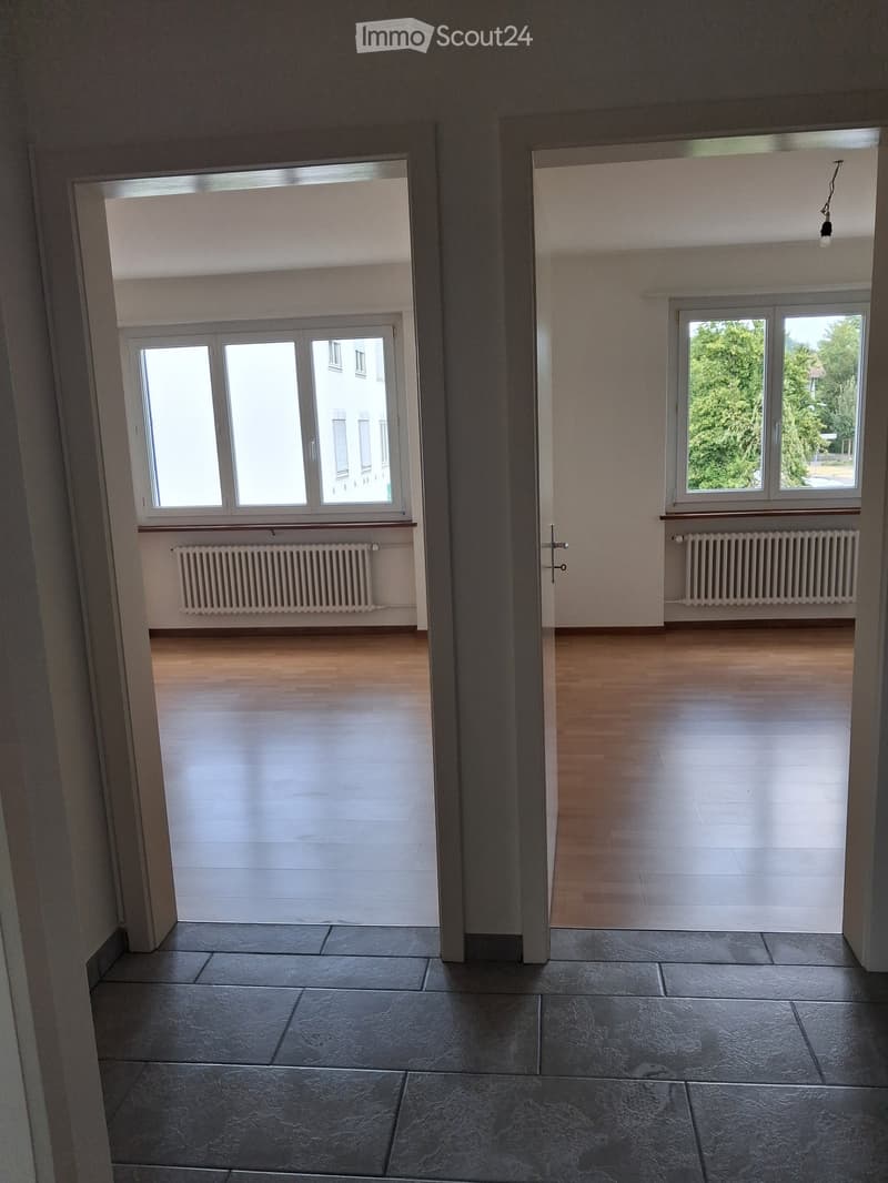 Empty room with wooden flooring, large windows, radiators, and a view of greenery outside. No furniture or equipment present.
