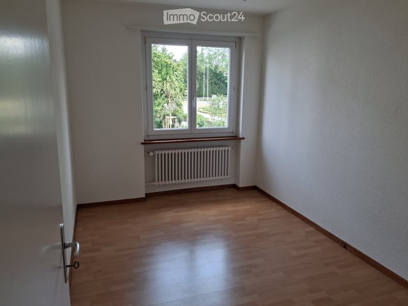 Empty room with wooden flooring, white walls, a window with a radiator below, and a door on the left side.