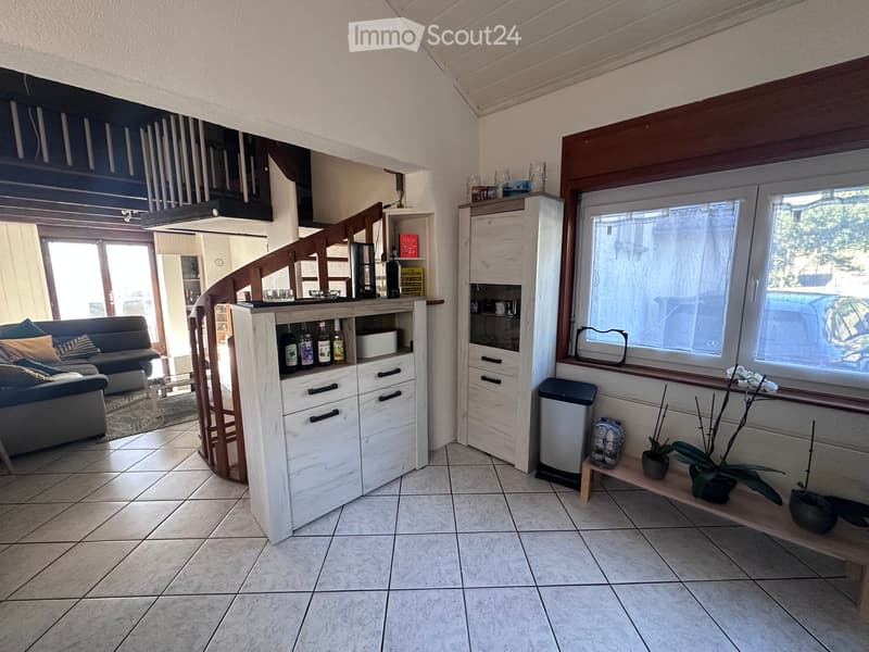 Living room with a sofa, coffee table, cabinet, bookshelf, potted plant, and trash bin. The room features large windows and a staircase leading to another level.