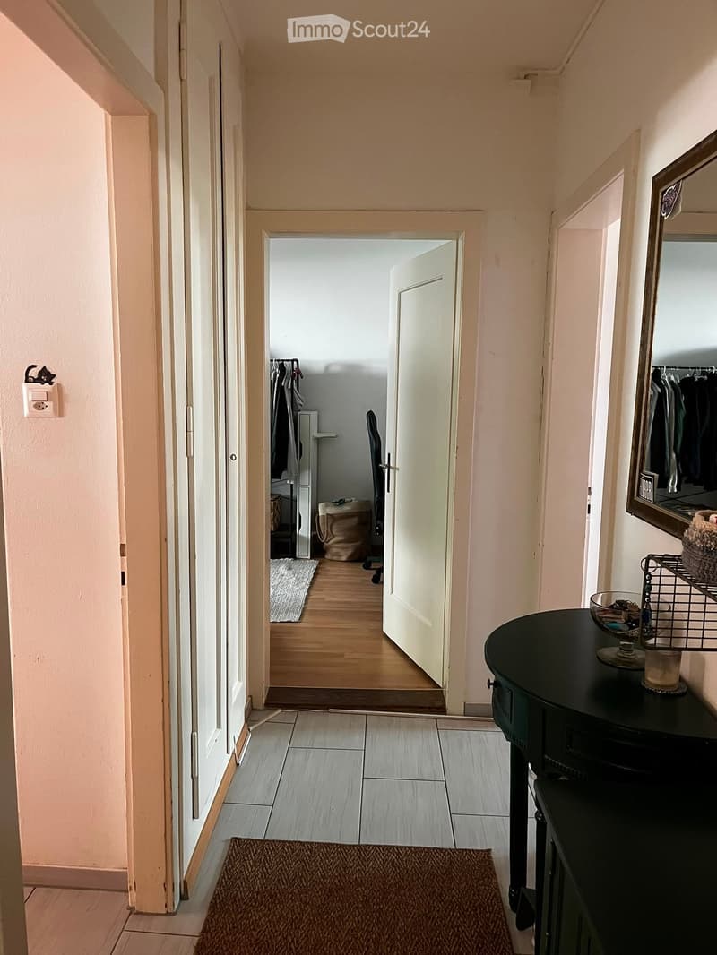 A narrow hallway with tiled flooring, light-colored walls, and several doors leading to other rooms. A small black table with a decorative bowl and a mirror is visible on the right side.