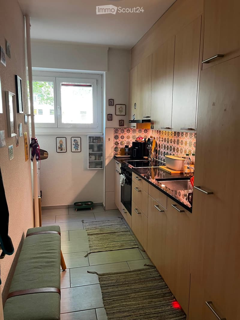 A modern kitchen with light wooden cabinets, a stainless steel sink, a refrigerator, and a small green stool near the entrance.
