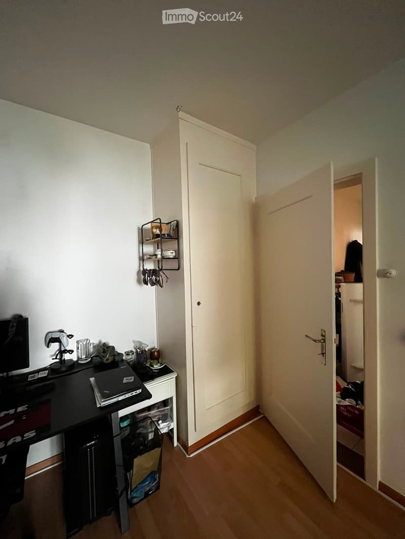 A small, furnished room with a wooden floor, a black table holding a laptop, screen, and various items, a small shelf with cups and other items, and an open door leading to another room.