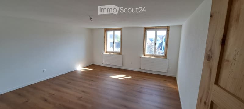 Empty room with wooden flooring, white walls, and two windows with wooden frames. Radiators are visible beneath the windows.