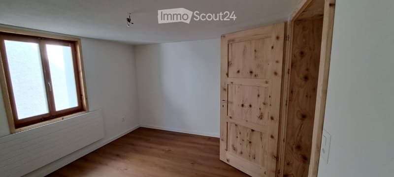Empty room with wooden flooring, white walls, a single window with wooden frame, and a partially open wooden door.