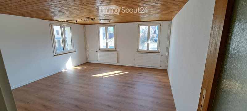 Empty room with wooden flooring, white walls, and three windows allowing natural light. The room features a wooden ceiling and built-in radiators along the walls.