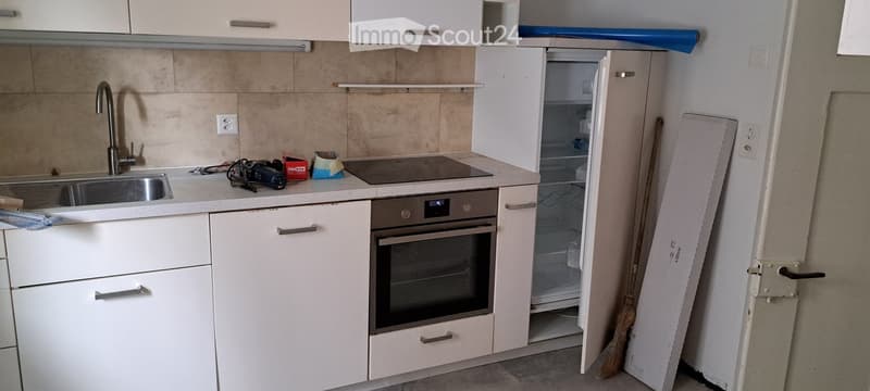 Modern kitchen with white cabinets, stainless steel sink, oven, and refrigerator. Countertop has a few items including a camera and some boxes.