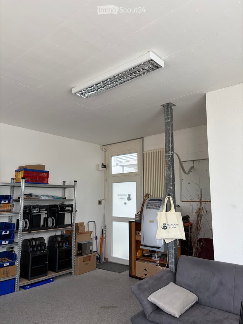 A furnished room with a grey sofa, a shelf filled with electronic equipment and storage boxes, an air conditioner, a handbag, and a radiator. The room has a grey carpeted floor and white walls.