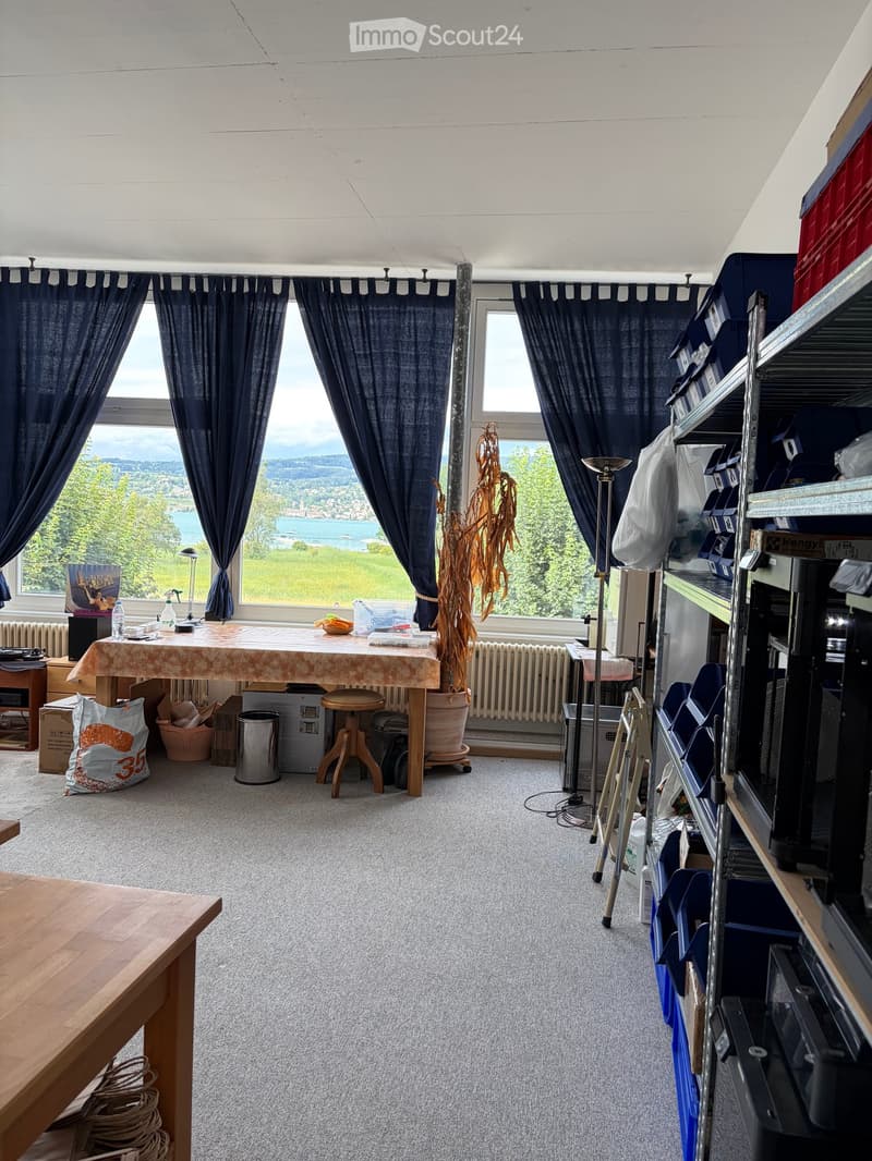 A furnished room with a large window offering a scenic view, a table with a patterned tablecloth, a stool, a shelf with various items, and a radiator beneath the window.