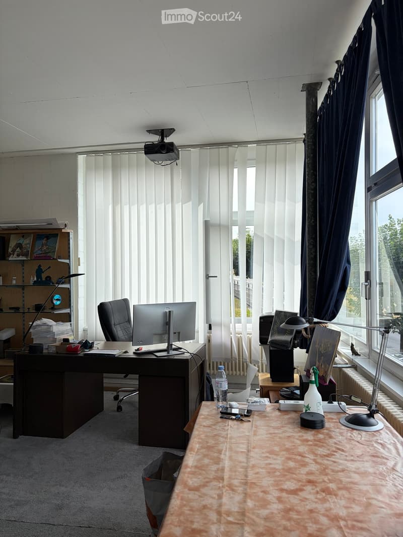 Well-lit office space with large windows, white blinds, and dark curtains. Features a desk with a computer monitor, keyboard, mouse, and various office supplies, along with a comfortable chair and a secondary table with additional items.