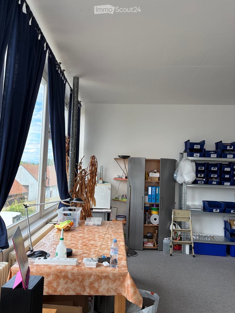 A furnished room with a table covered in a patterned cloth, a laptop, a bottle, a lamp, a refrigerator, shelves with storage boxes, and a stool near the window with a view of the outside.