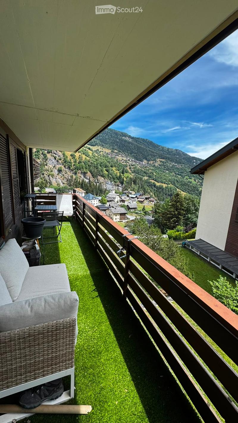 Balcony with green artificial turf, wooden railing, and scenic mountain view. Features a wicker sofa, small table, and potted plant.