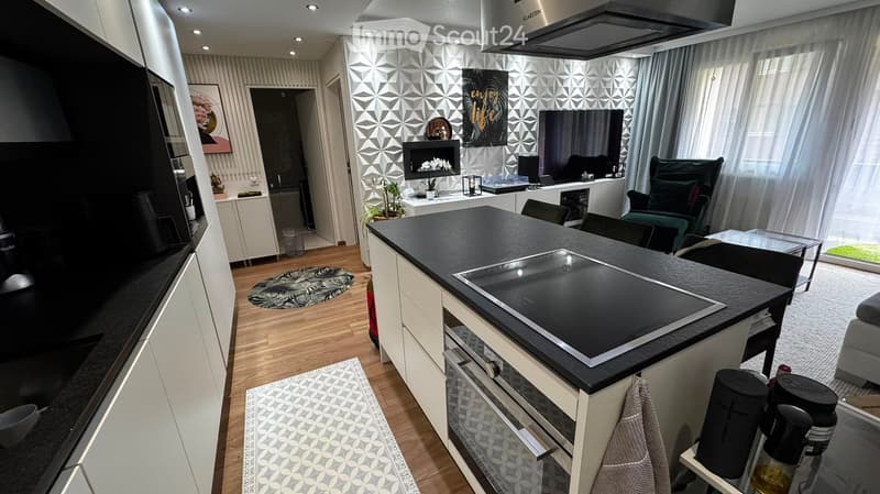 Modern open-plan kitchen and living area with a central island, induction cooktop, sleek cabinets, and a cozy living space featuring a sofa, TV, and fireplace.
