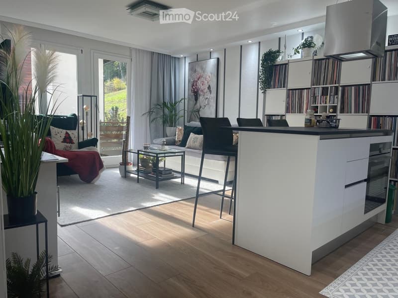 Modern living room with a white kitchen island, sofa, coffee table, and bookshelf. Large windows provide natural light, and the space is decorated with plants and artwork.
