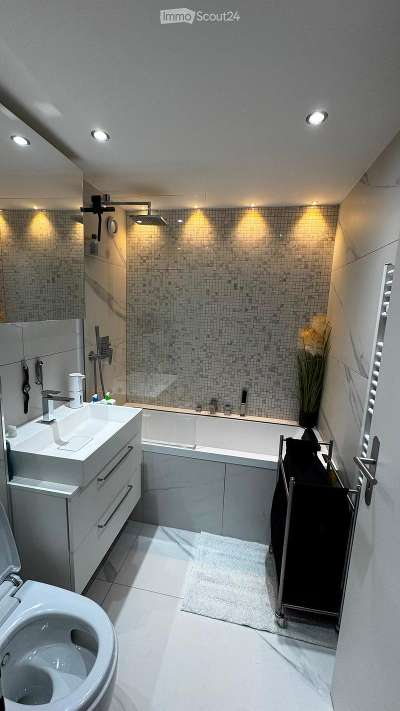 Modern bathroom with a white sink, toilet, bathtub, and shower area. Features include a towel rack, a small plant, and recessed lighting.