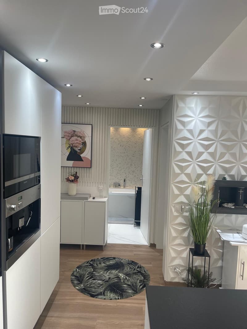 Modern kitchen with white cabinets, stainless steel oven, round patterned rug, and decorative elements including a vase with flowers and a geometric wall design.