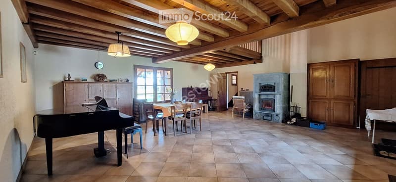 A spacious room with exposed wooden beams, a grand piano, a dining table with chairs, a fireplace, and a cabinet.