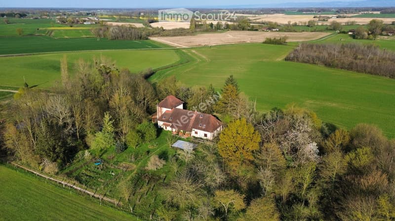 Aerial view of a single-story house with a red-tiled roof, surrounded by trees and greenery, located in a rural area with open fields in the background.