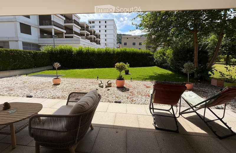 A modern outdoor patio with a wicker sofa, wooden table, and lounge chairs, surrounded by potted plants and a well-maintained lawn.
