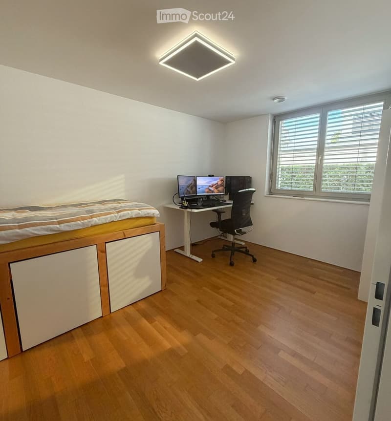 Modern bedroom with a lofted bed, desk with computer and dual monitors, ergonomic chair, and large window with blinds.