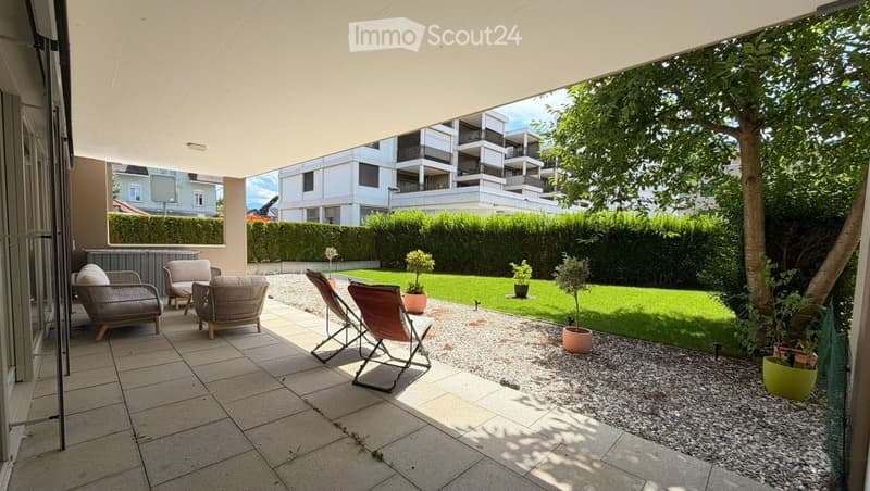 A modern outdoor patio with tiled flooring, lounge chairs, and potted plants, overlooking a well-maintained lawn and modern apartment buildings.