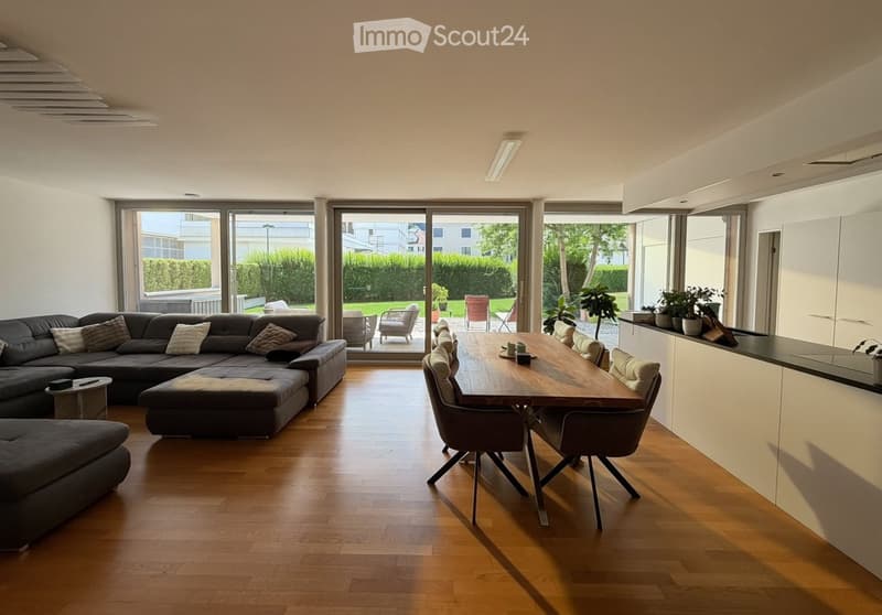 Bright living and dining area with wooden flooring, large sliding glass doors leading to a patio, a dark sectional sofa, a wooden dining table with chairs, and a modern kitchen counter with potted plants.