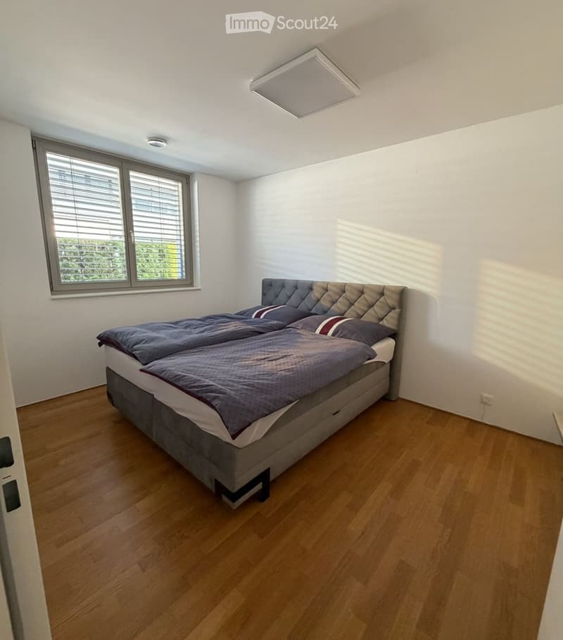 Modern bedroom with a large double bed, wooden flooring, and large window with blinds. The bed features a tufted headboard and is made up with blue bedding and pillows.