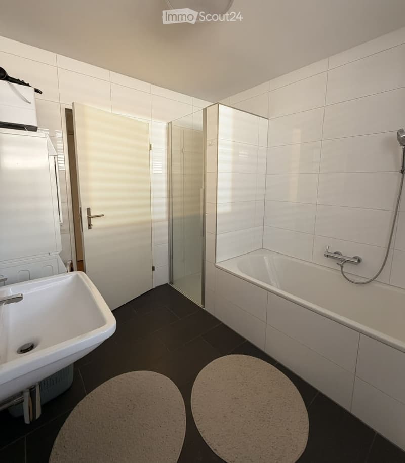 Modern bathroom with white tiles, a large sink, a bathtub with a glass shower enclosure, and two round rugs on the dark tiled floor.
