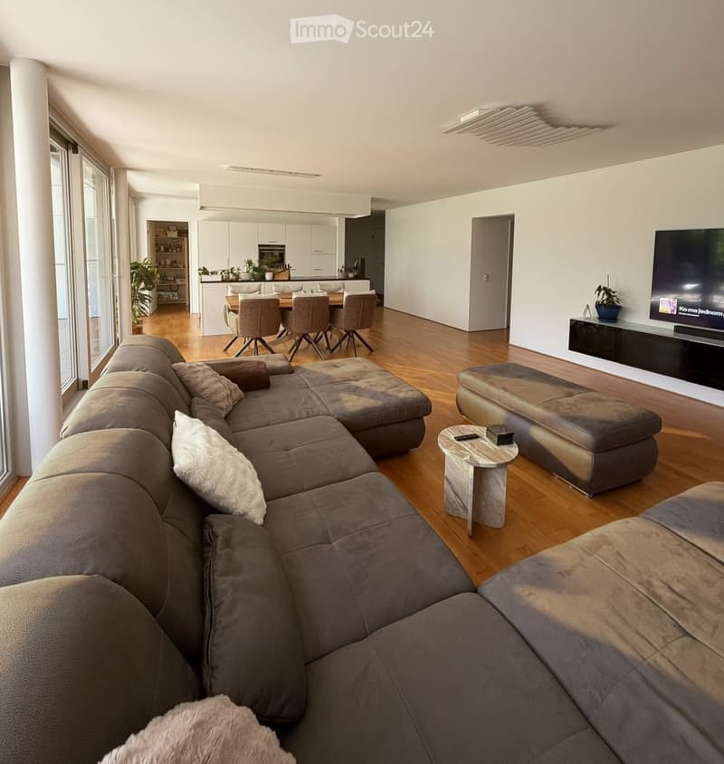 Modern living room with a large sectional sofa, coffee table, ottoman, and a TV mounted on the wall. The space is open-plan, connecting to a dining area with a table and chairs, and a kitchen in the background.