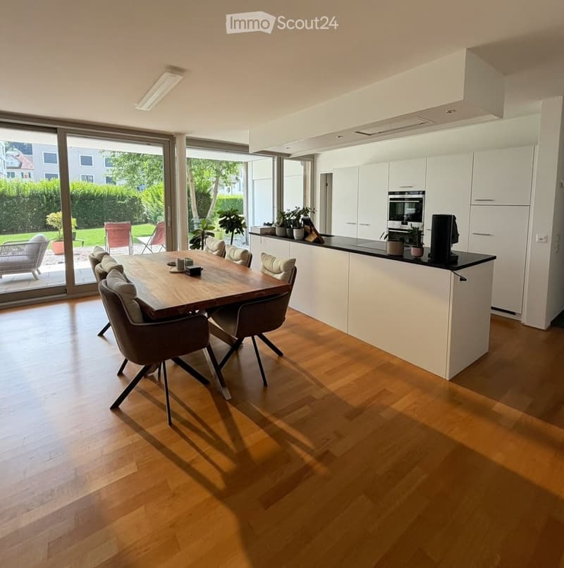 Open-plan kitchen and dining area with light wood flooring, white cabinets, a wooden dining table with chairs, and a modern oven. Large sliding glass doors lead to a garden with outdoor seating.