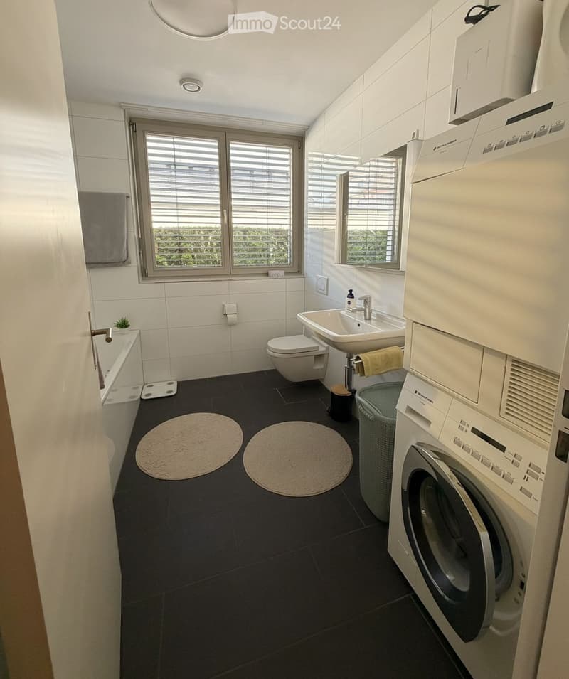 Modern bathroom with white tiles, black floor, bathtub, toilet, sink, washing machine, and two round rugs.