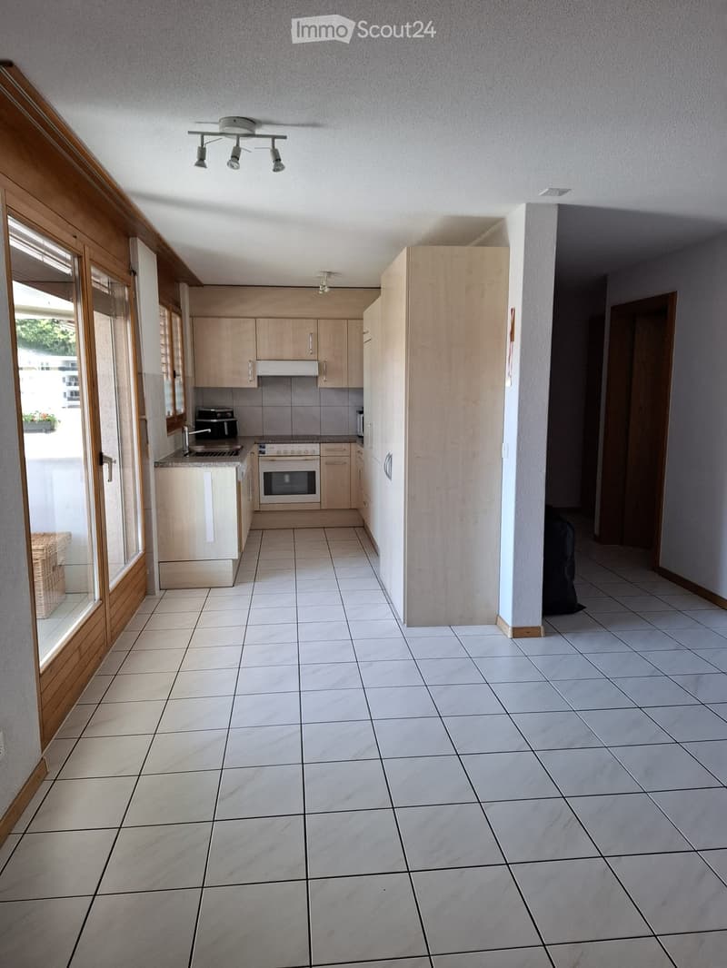 Bright kitchen with white tiled floor, light wooden cabinets, and stainless steel appliances including an oven and fridge. A large window with wooden frame provides natural light.