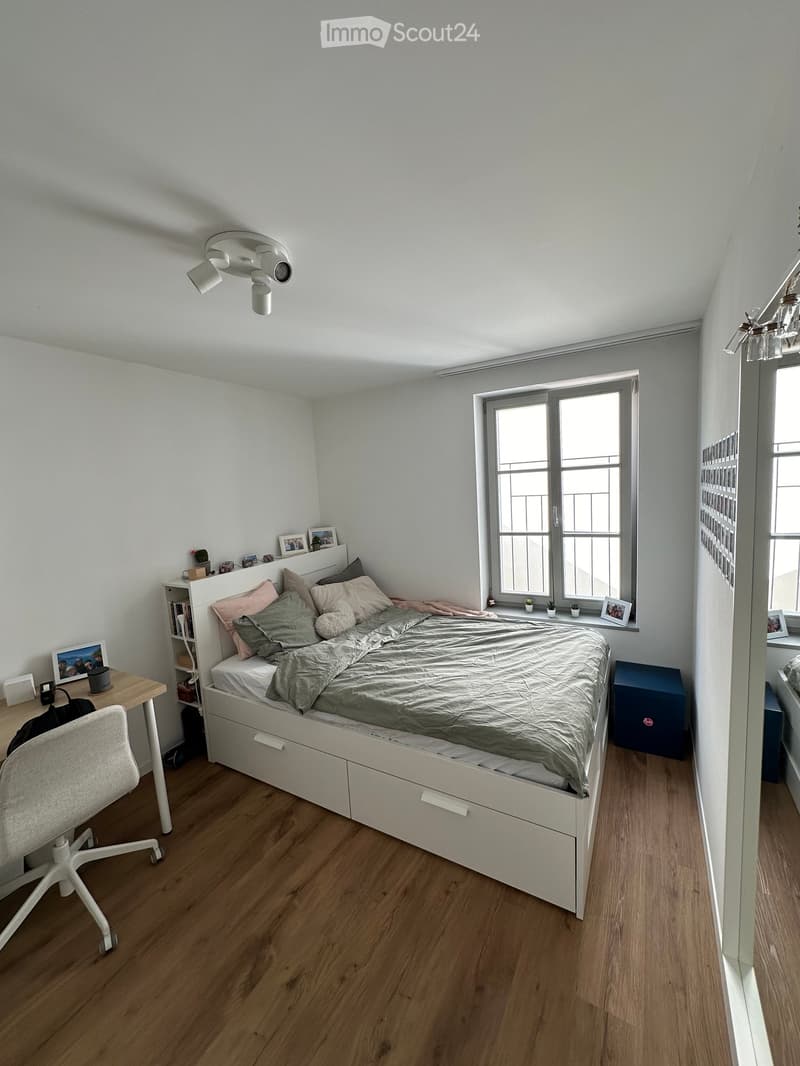Cozy bedroom with a white bed, wooden floor, desk, chair, and shelves. The bed has multiple pillows and a duvet, and there are picture frames on the shelves and windowsill.