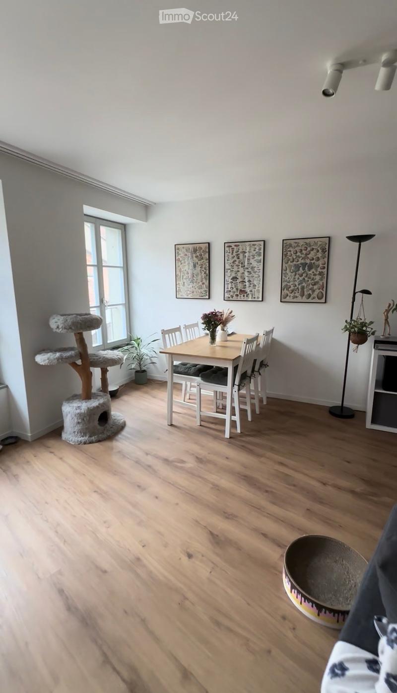 Bright dining area with wooden floor, white walls, and large window. Includes a dining table with chairs, a cat tree, a vase with flowers, a potted plant, a floor lamp, and several picture frames on the wall.