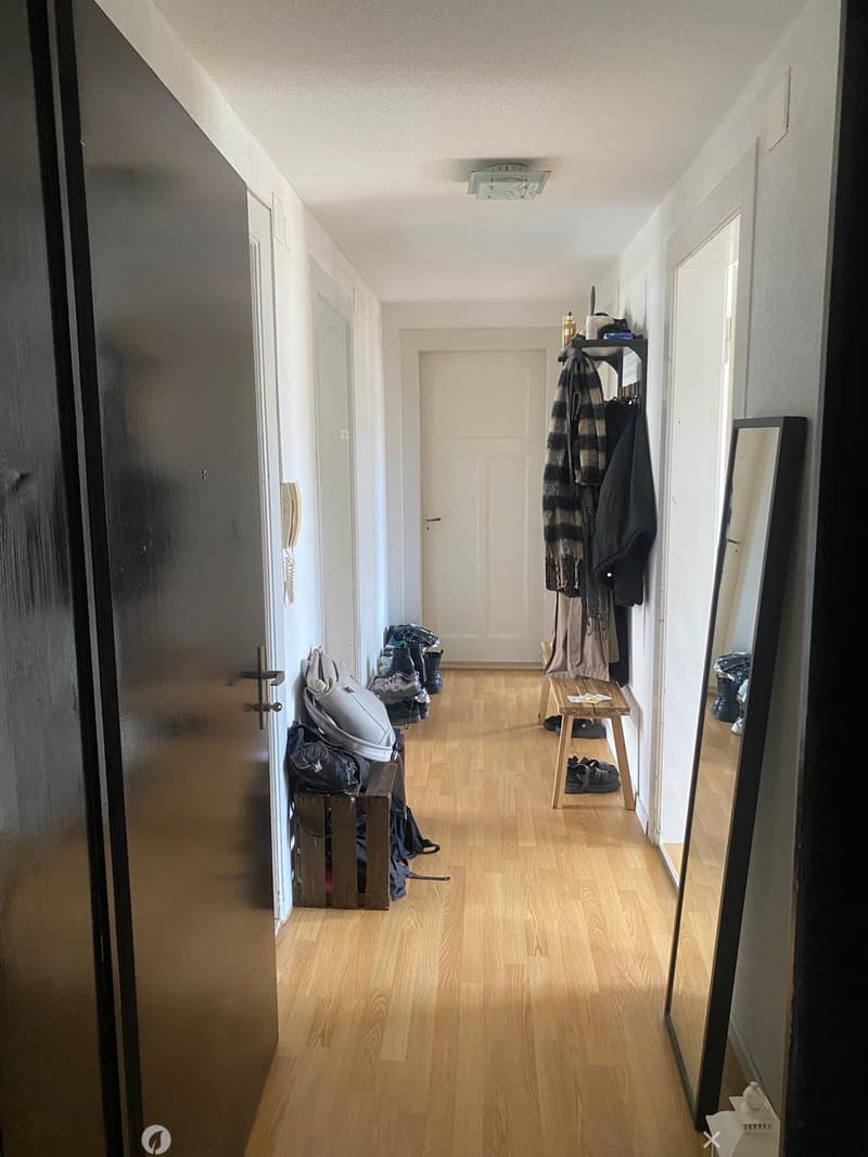 A narrow hallway with wooden flooring, white walls, and several doors. There are coats and shoes placed on small benches on either side.