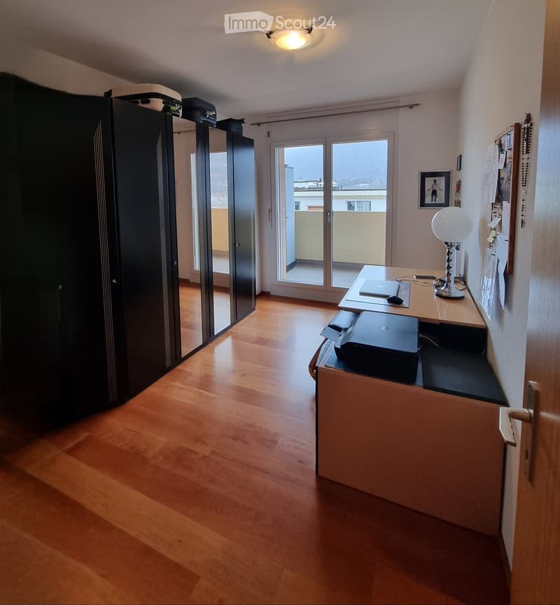 wooden floor, large wardrobe, desk with computer, lamp, sliding glass door to balcony
