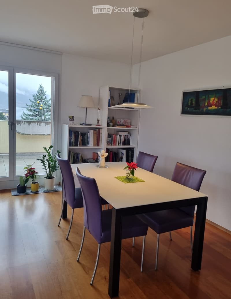 Dining room with a white table, four purple chairs, a white shelf with books, a lamp, a picture frame, and a glass door leading to a balcony with a view of a tree and a building.