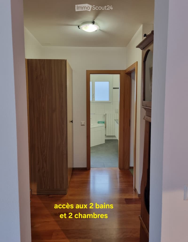 Empty hallway with wooden floor, white walls, wooden doors, and a ceiling light. The hallway leads to a bathroom and two bedrooms.