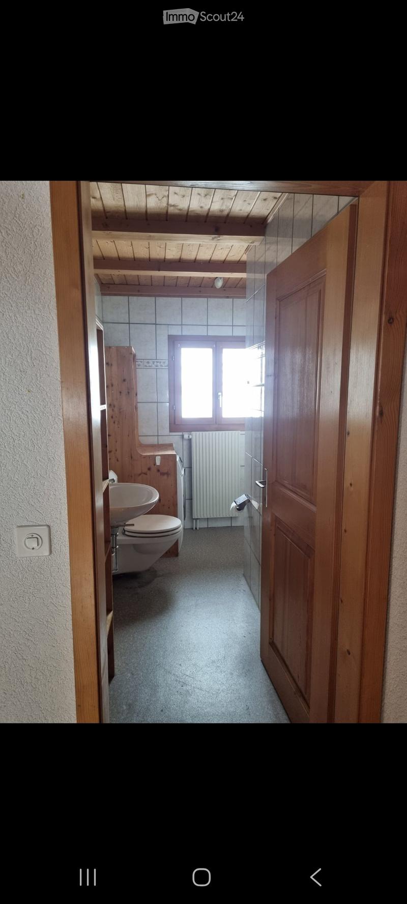 Bathroom with wooden ceiling, tiled walls, toilet, sink, and radiator. The space is connected to a hallway with wooden doors and a window providing natural light.