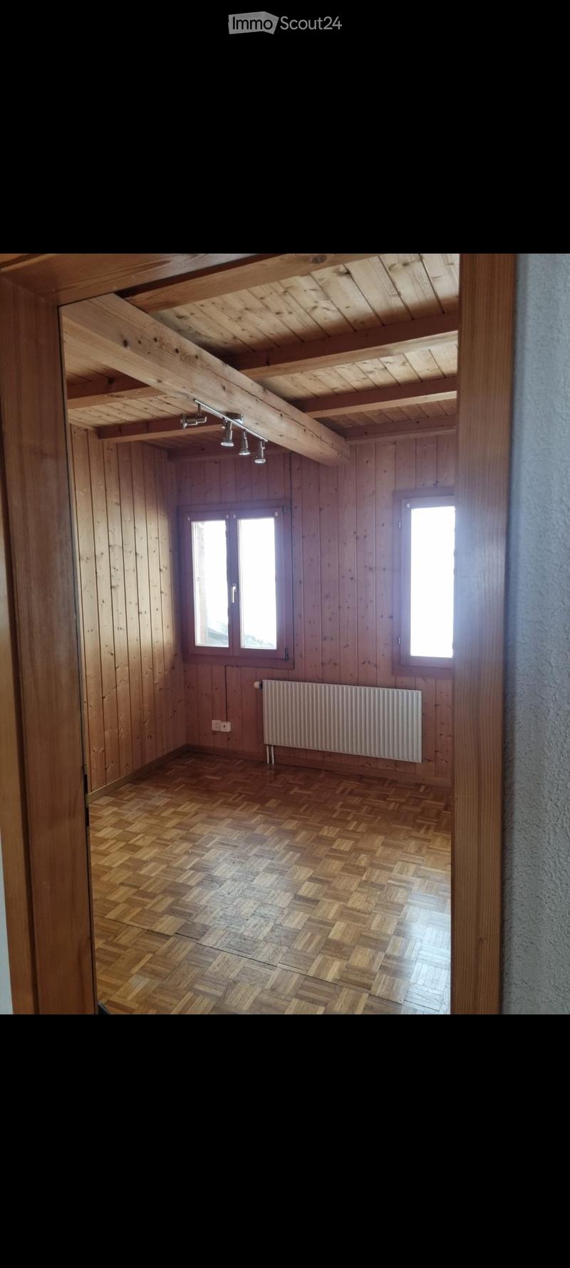 Empty room with wooden walls, herringbone wooden floor, exposed wooden beams on the ceiling, and a radiator on one wall.