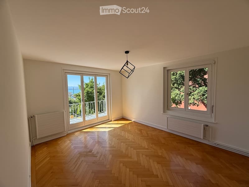 Empty room with wooden herringbone flooring, large windows, and a sliding glass door leading to a balcony.