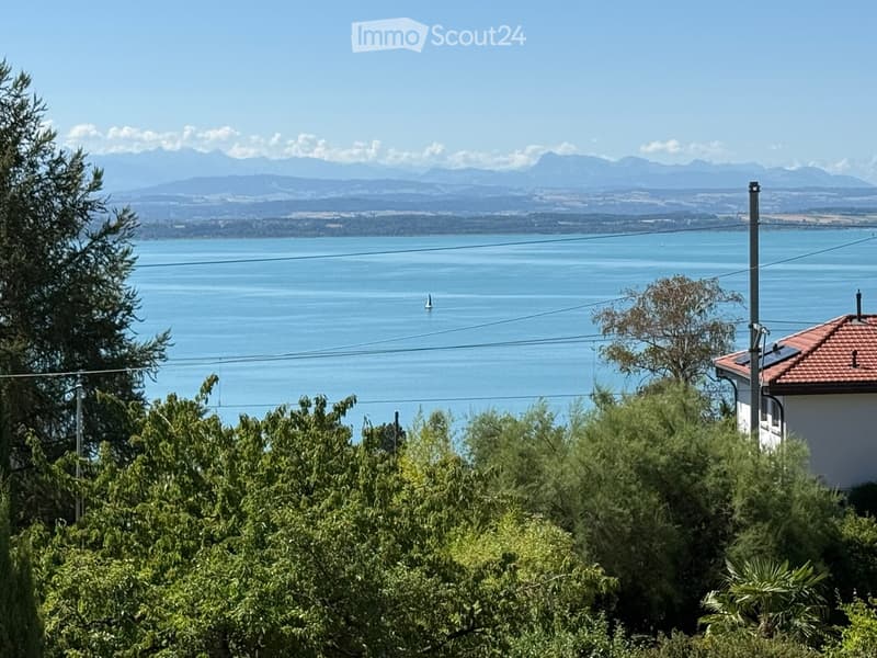 A scenic view of a lake with distant mountains, surrounded by lush greenery and a small house with a red-tiled roof on the right side.