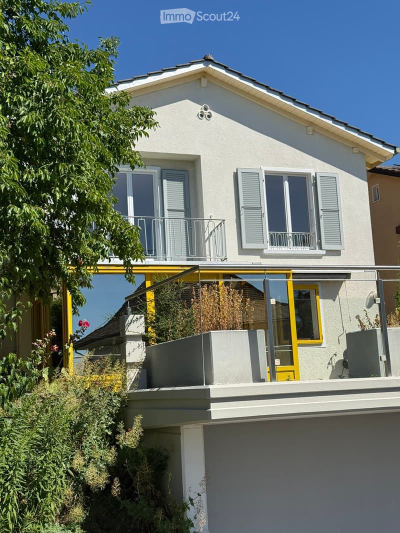 Two-story house with white exterior, grey shutters, and a balcony on the upper floor. The lower level features a modern glass-enclosed terrace with yellow framing and some potted plants.