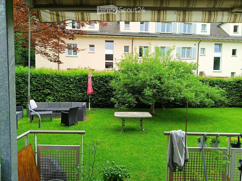 Garden with green lawn, patio furniture including a sofa, table, and chairs, surrounded by hedges and a building in the background.