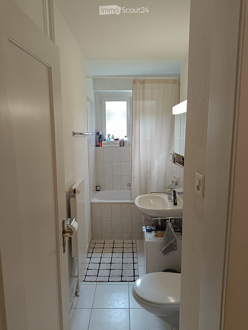 Modern bathroom with white tiles, bathtub, sink, toilet, and a small window above the bathtub. Includes a shower curtain, towel, and a small cabinet with toiletries.