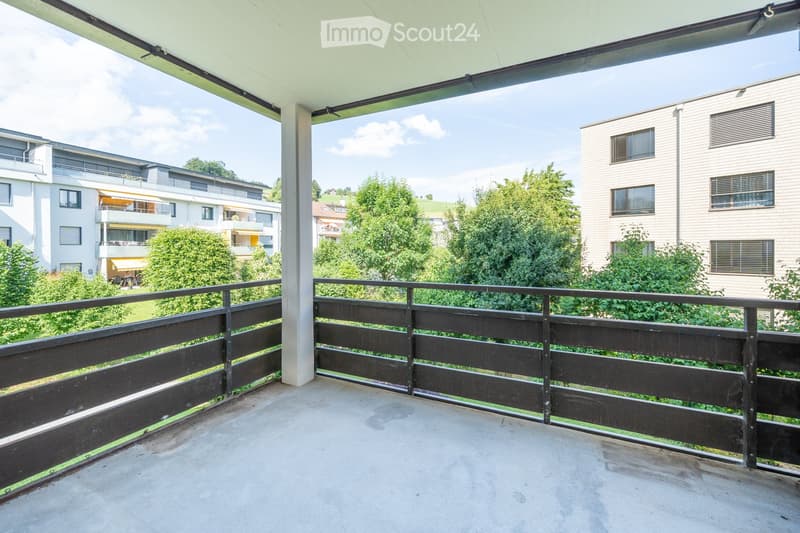 Balcony with wooden railing, concrete floor, and view of surrounding greenery and buildings.