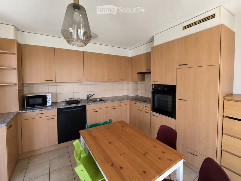 Modern kitchen with light wooden cabinets, granite countertops, and stainless steel appliances including a microwave, dishwasher, and oven. A wooden dining table with green and purple chairs is positioned in the center.