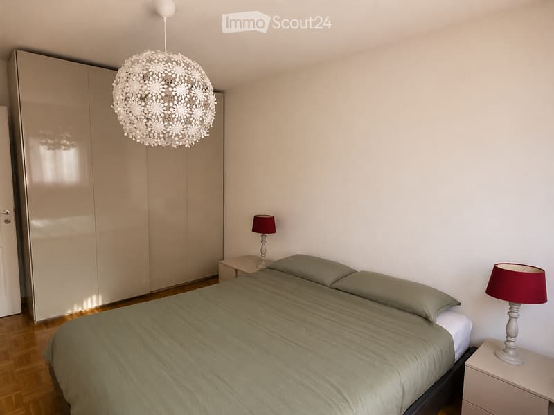 Bedroom with a double bed, nightstands, lamps, and a large wardrobe. The room features wooden flooring and white walls.