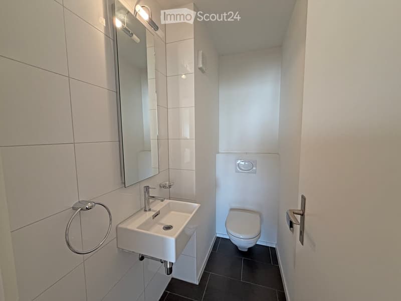 Modern, compact bathroom with white tiles, a wall-mounted sink, a toilet, a large mirror, and a towel ring.
